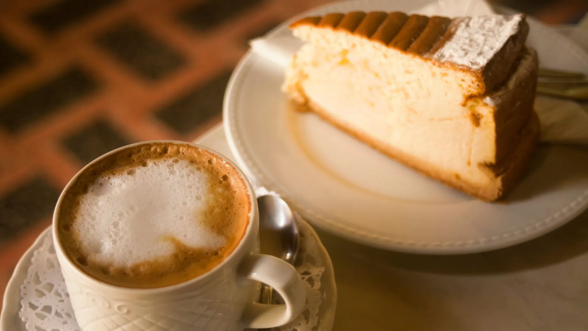 Slice of cheesecake on a white plate next to a frothy cappuccino in a white cup
