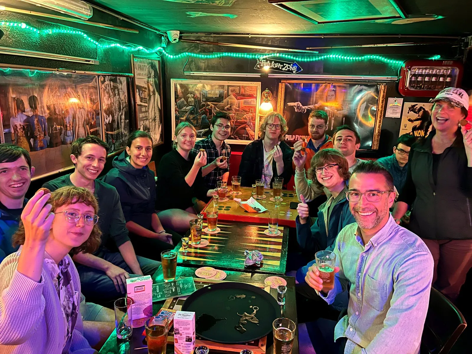A large group raising drinks around tables in a lively neon-lit Amsterdam bar