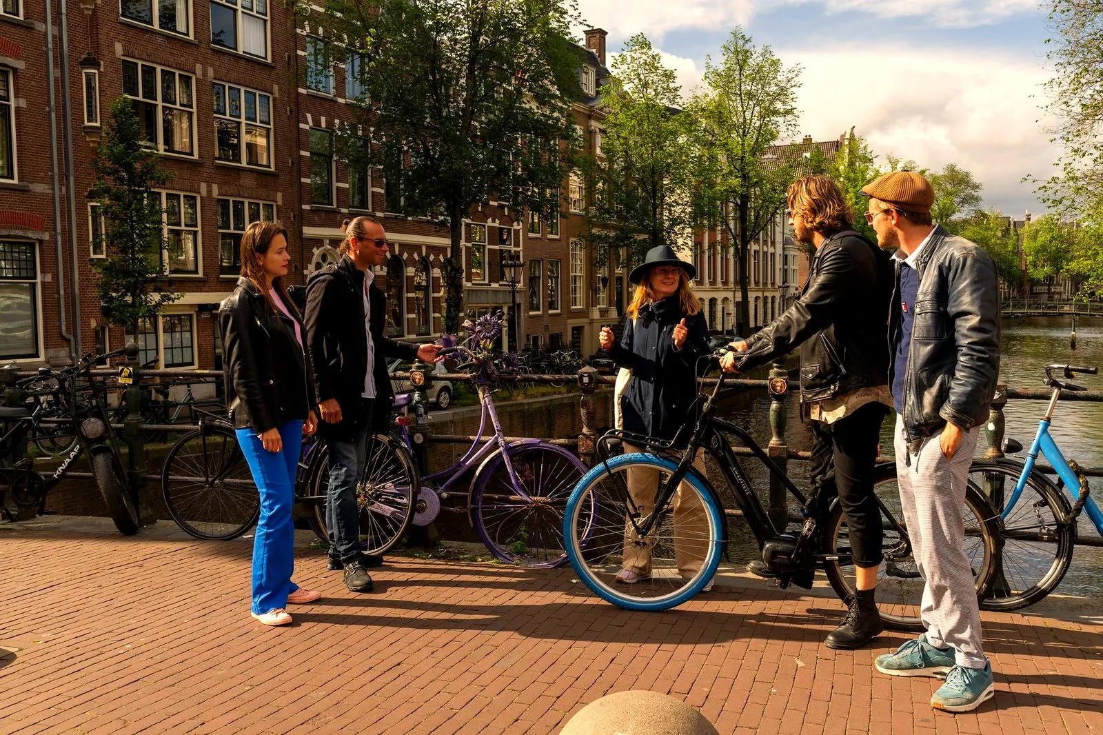 Small group with bicycles chatting on a sunny Amsterdam canal bridge between old brick canal houses