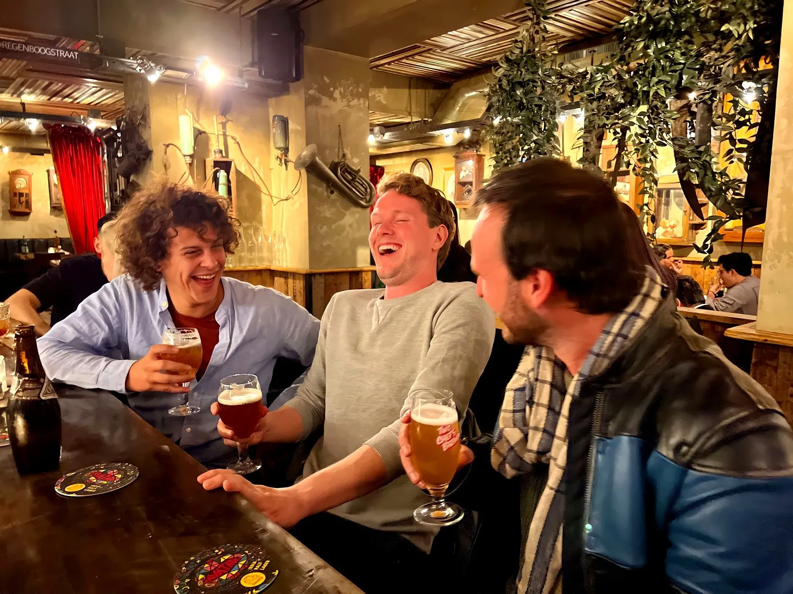 Three friends laughing over glasses of beer in a cosy Amsterdam brown bar