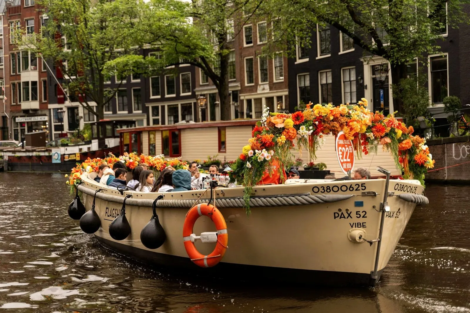 An open canal boat decorated with orange flowers carrying guests past Amsterdam canal houses