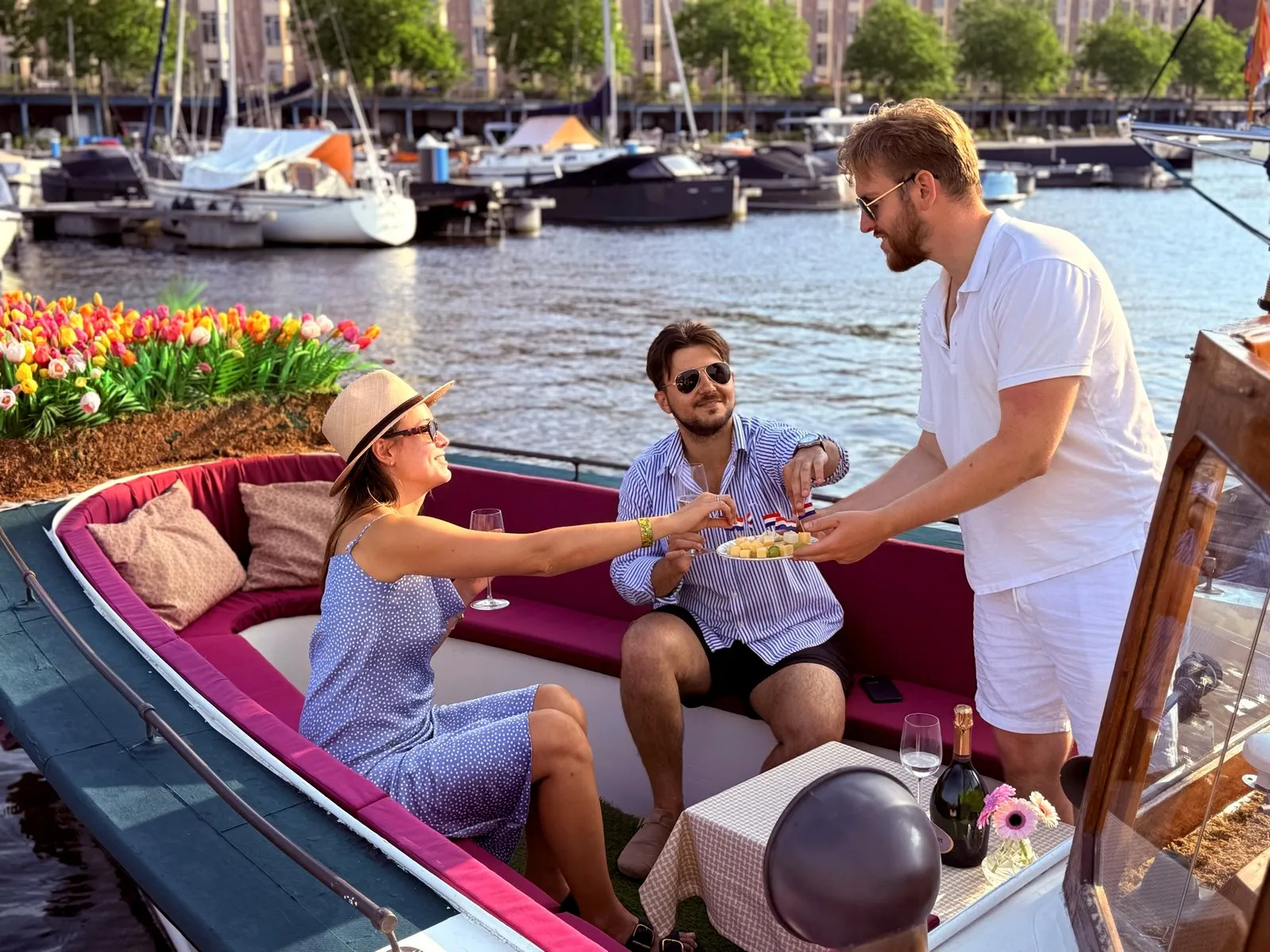 A host on a canal boat serving snacks to two seated guests with drinks, tulips behind