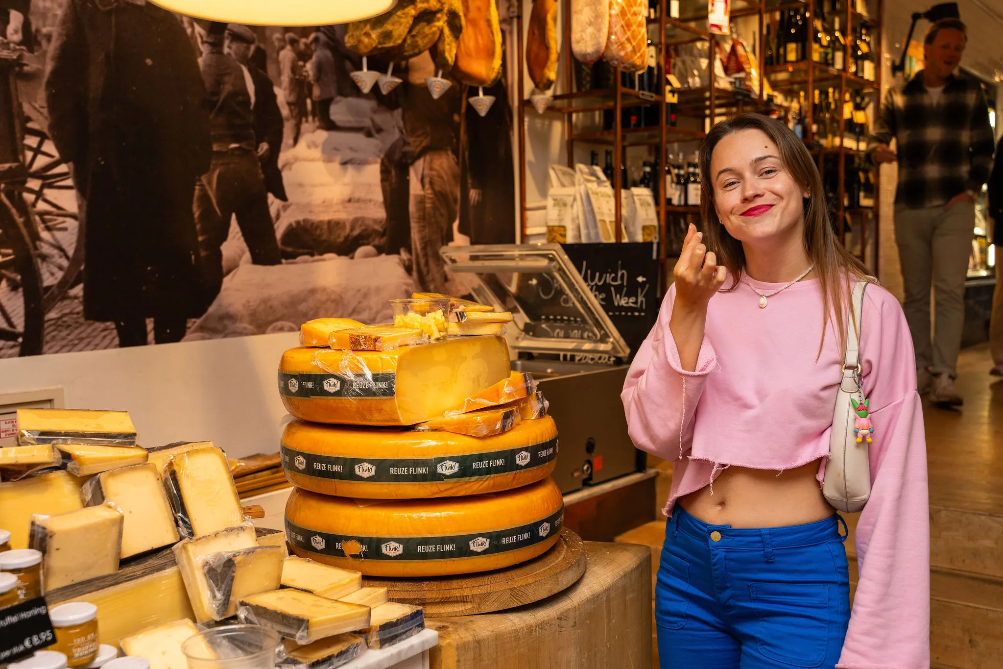 Woman in a pink top making a chef's kiss beside stacked Gouda cheese wheels in an Amsterdam cheese shop