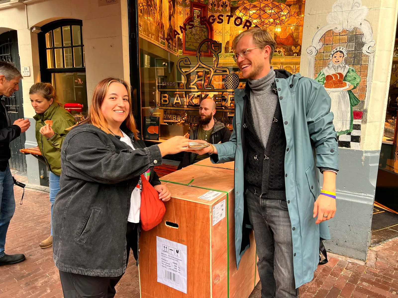 A tasting is handed to a smiling guest in a rain jacket outside an Amsterdam bakery