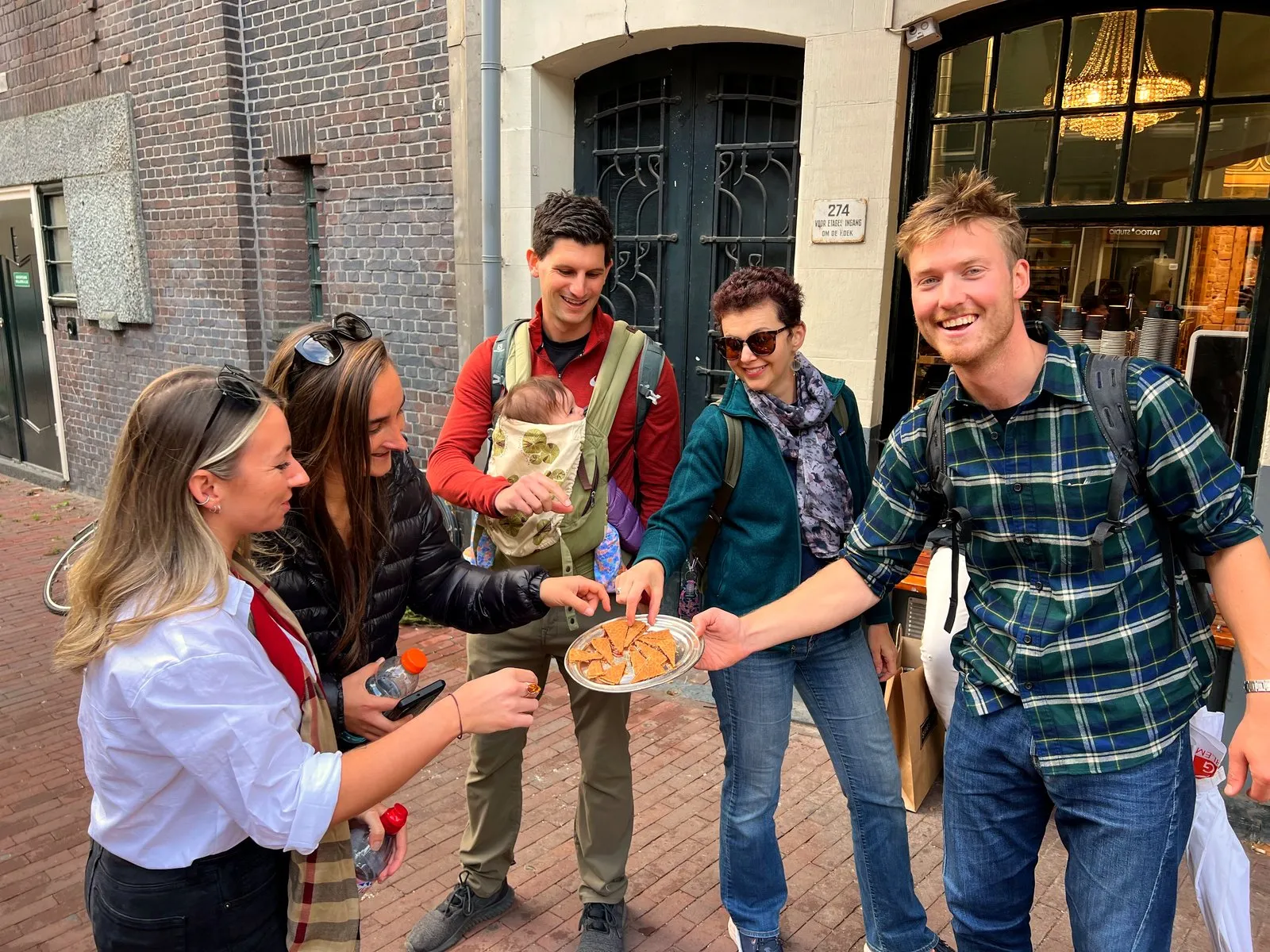 A food tour group on an Amsterdam street reaches for stroopwafel pieces on a plate, one guest carrying a baby in a front carrier