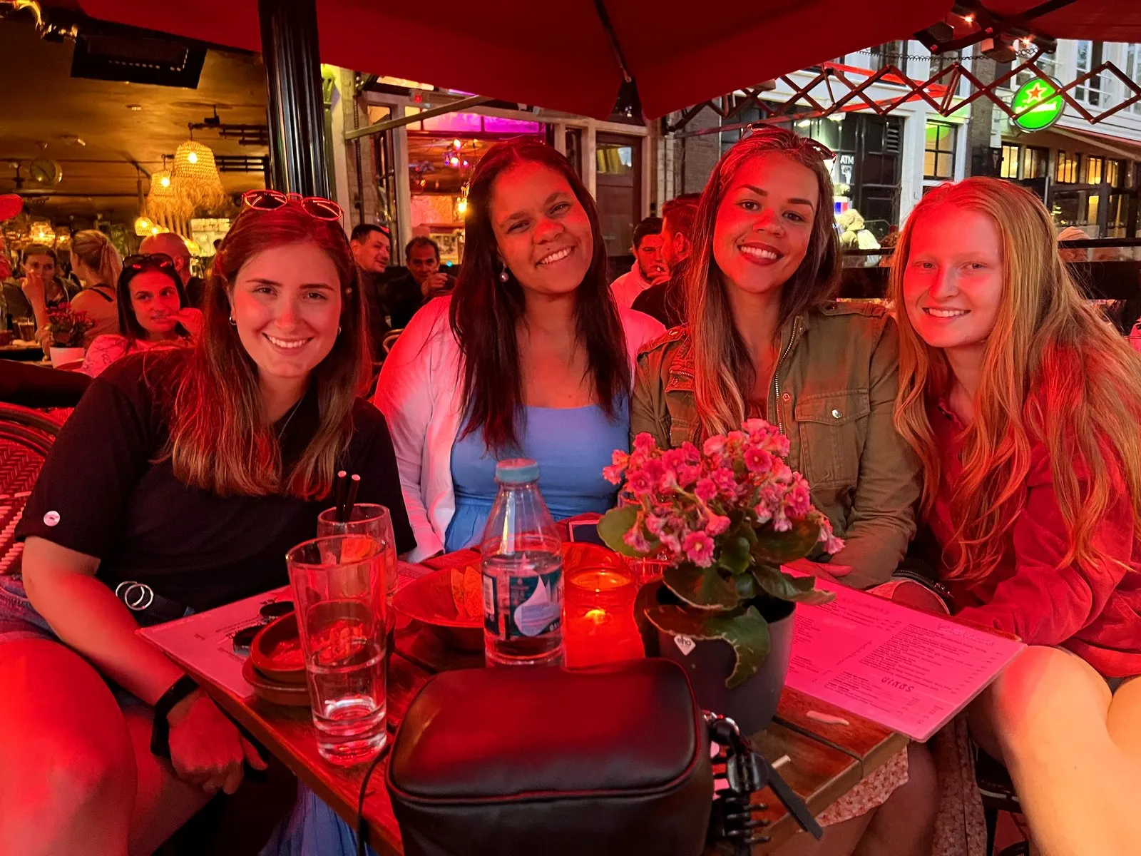 Four young women smiling at a red-lit bar table in Amsterdam at night