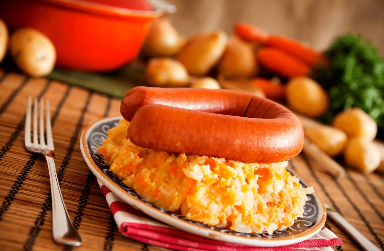 Plate of hutspot mash with a whole smoked rookworst on top, potatoes and carrots on the table behind it