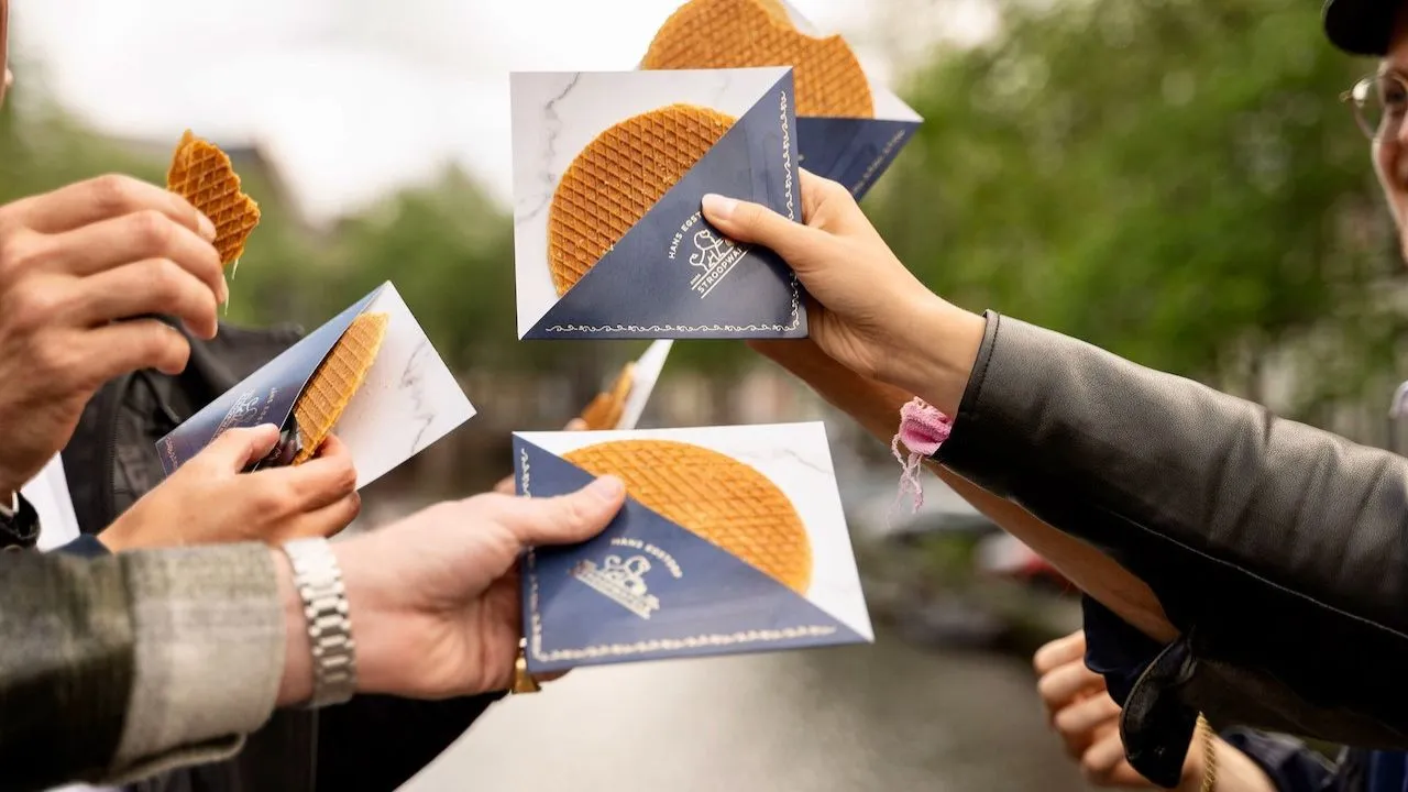 Several hands holding fresh stroopwafels in blue paper sleeves outdoors by a canal