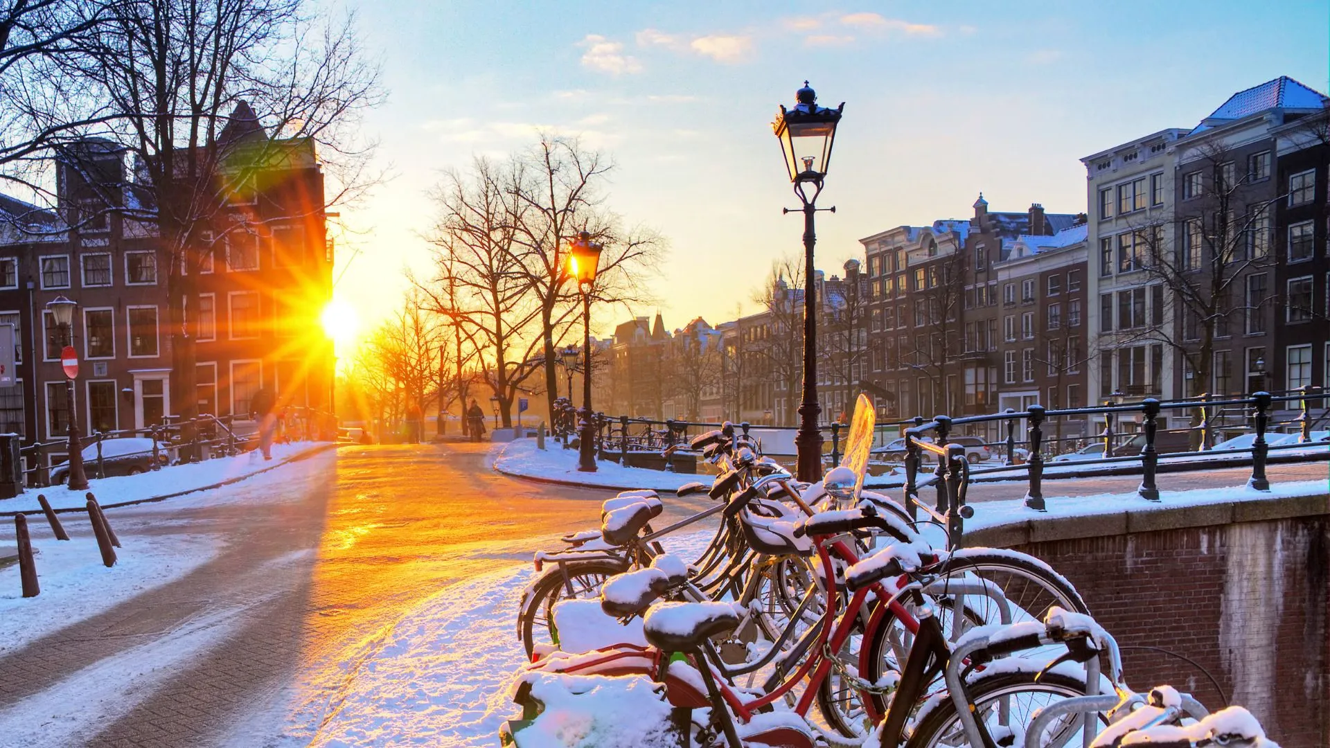 Snow-covered bicycles parked on an Amsterdam canal bridge at sunrise, with lampposts and bare trees in golden light