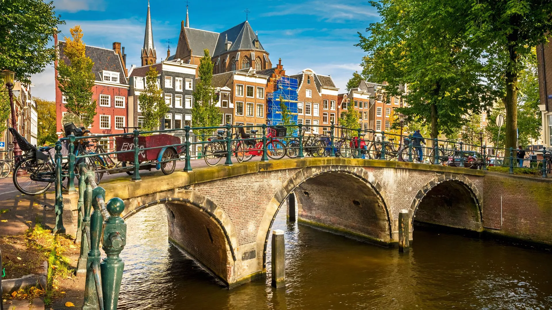 Arched stone bridge over an Amsterdam canal with cyclists and parked bikes on top and church spires behind