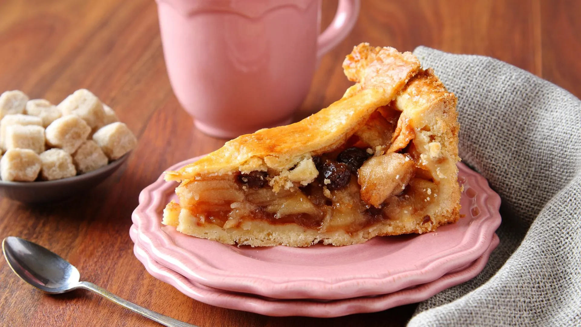 Slice of Dutch apple pie with raisins and a golden lattice crust on a pink plate, sugar cubes and a spoon nearby