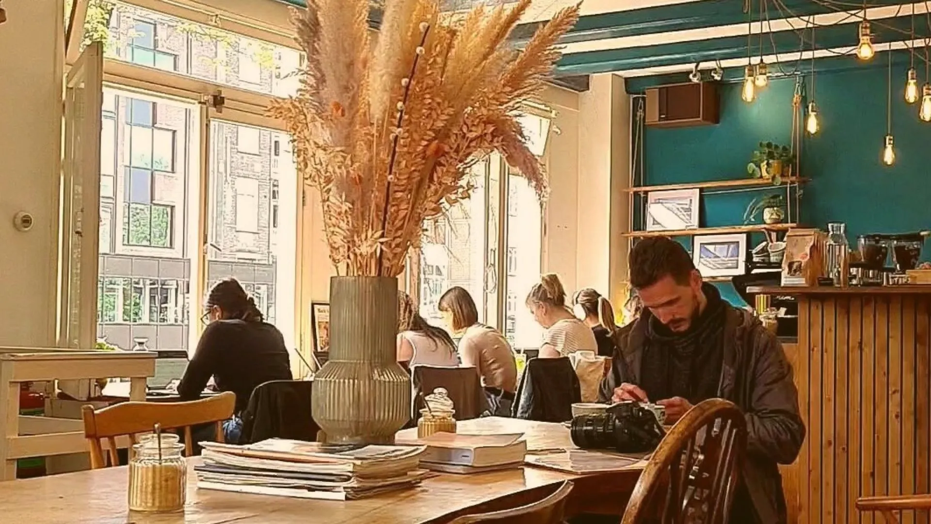 Cozy cafe interior with a teal ceiling, hanging bulbs, dried pampas grass in a vase and people reading at wooden tables