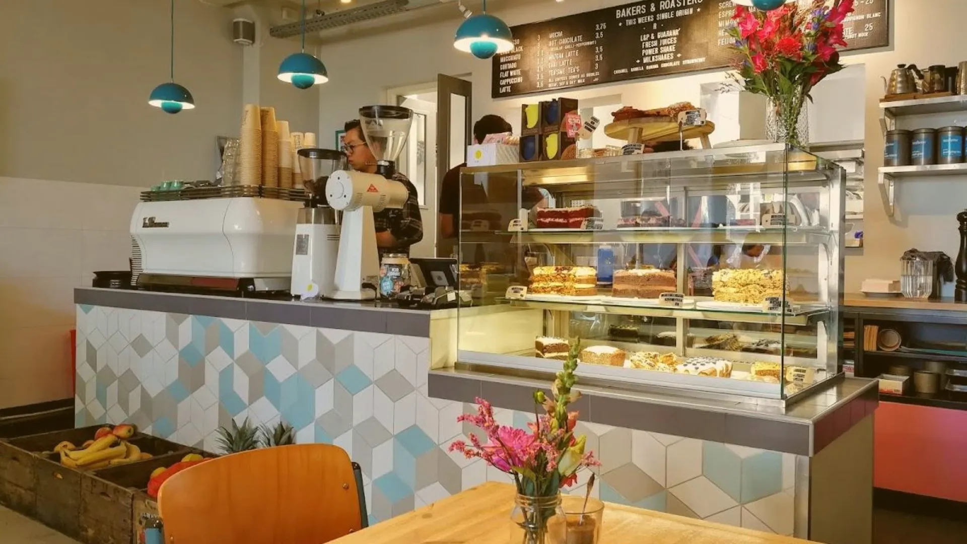 Cafe counter with an espresso machine, a glass display case full of cakes and banana bread, and staff at work behind