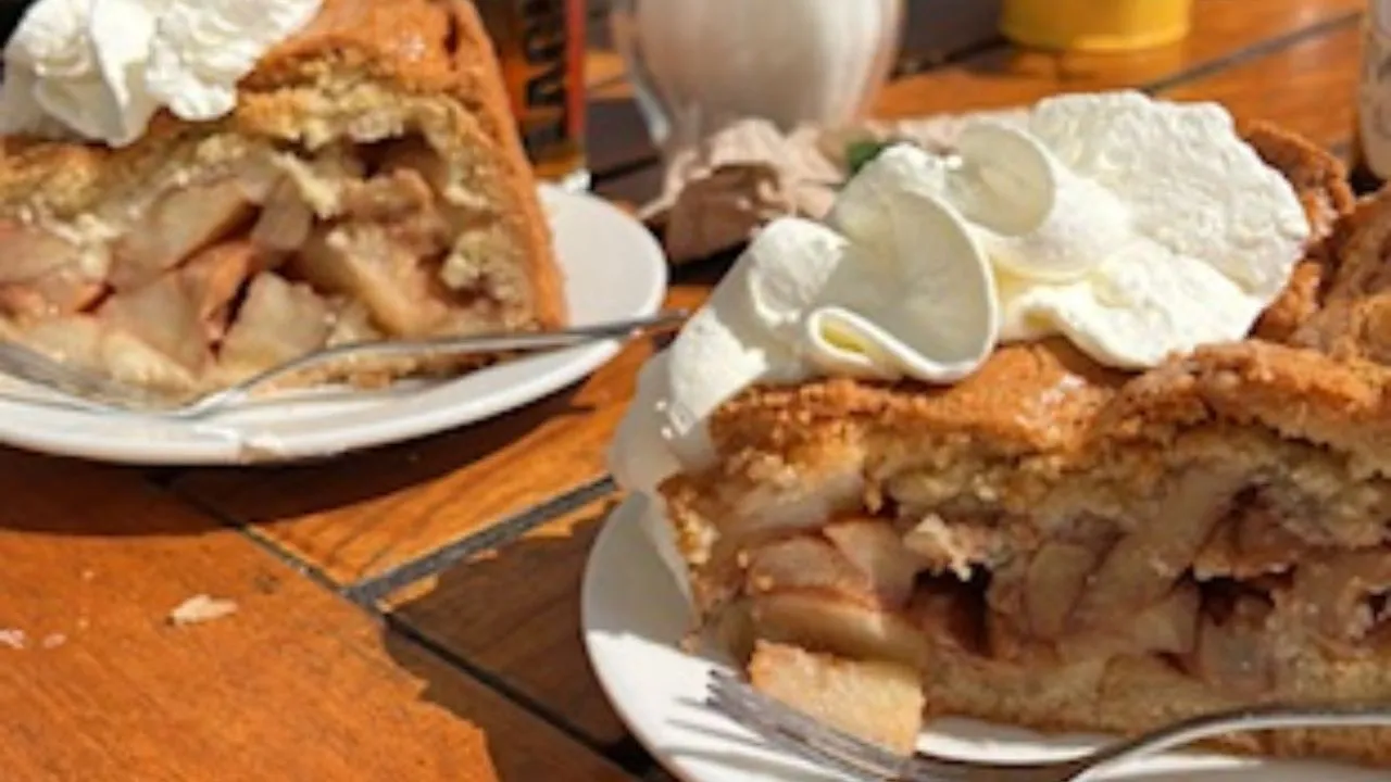 Fresh Dutch apple pie slices topped with whipped cream on a sunny outdoor café table in Amsterdam, with drinks and flowers in the background.