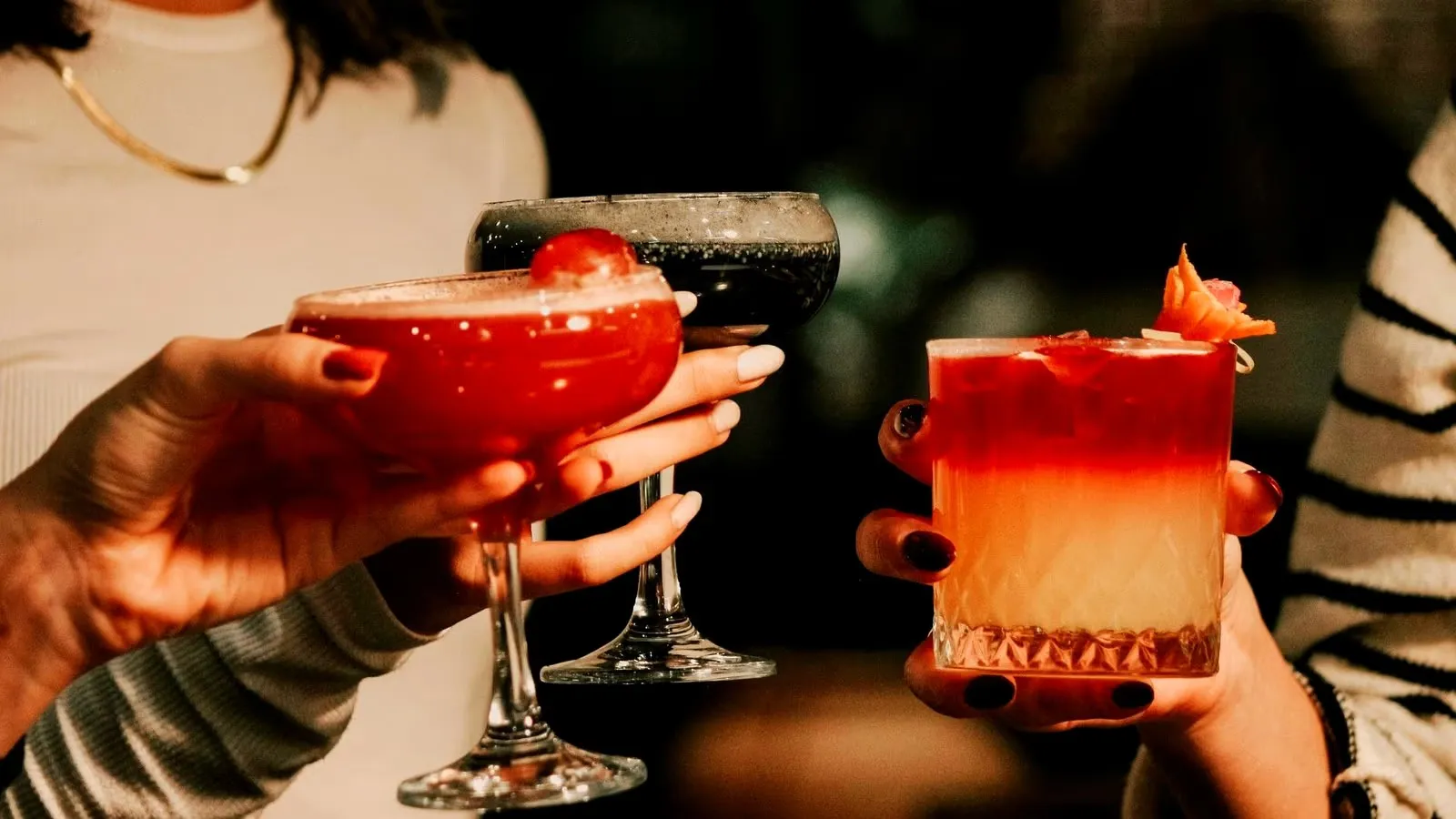 Three hands clinking colorful cocktails in a dimly lit Amsterdam bar