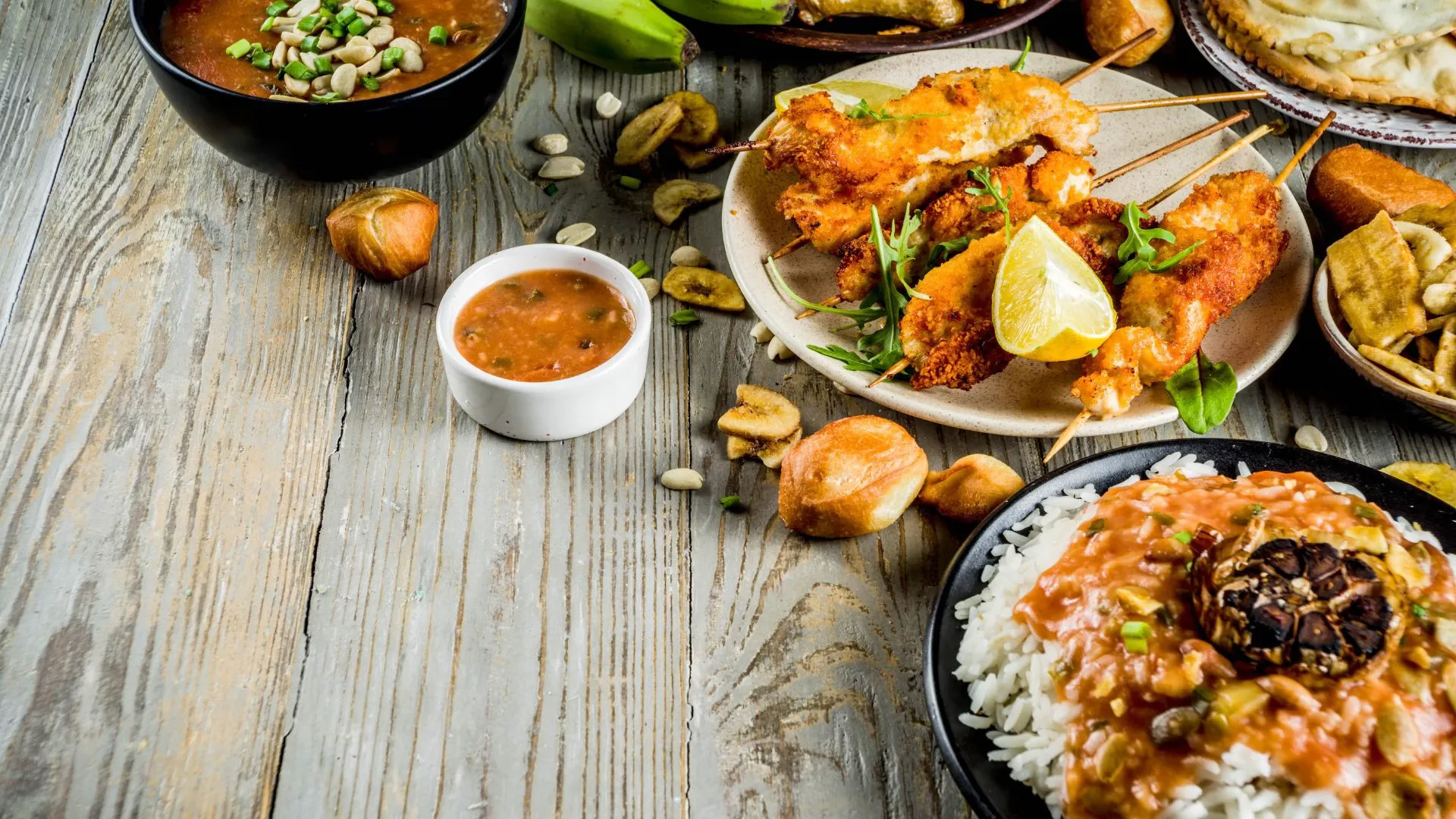 Wooden table spread with West African dishes including chicken skewers with lime, rice topped with stew, fried plantains and a bowl of peanut soup