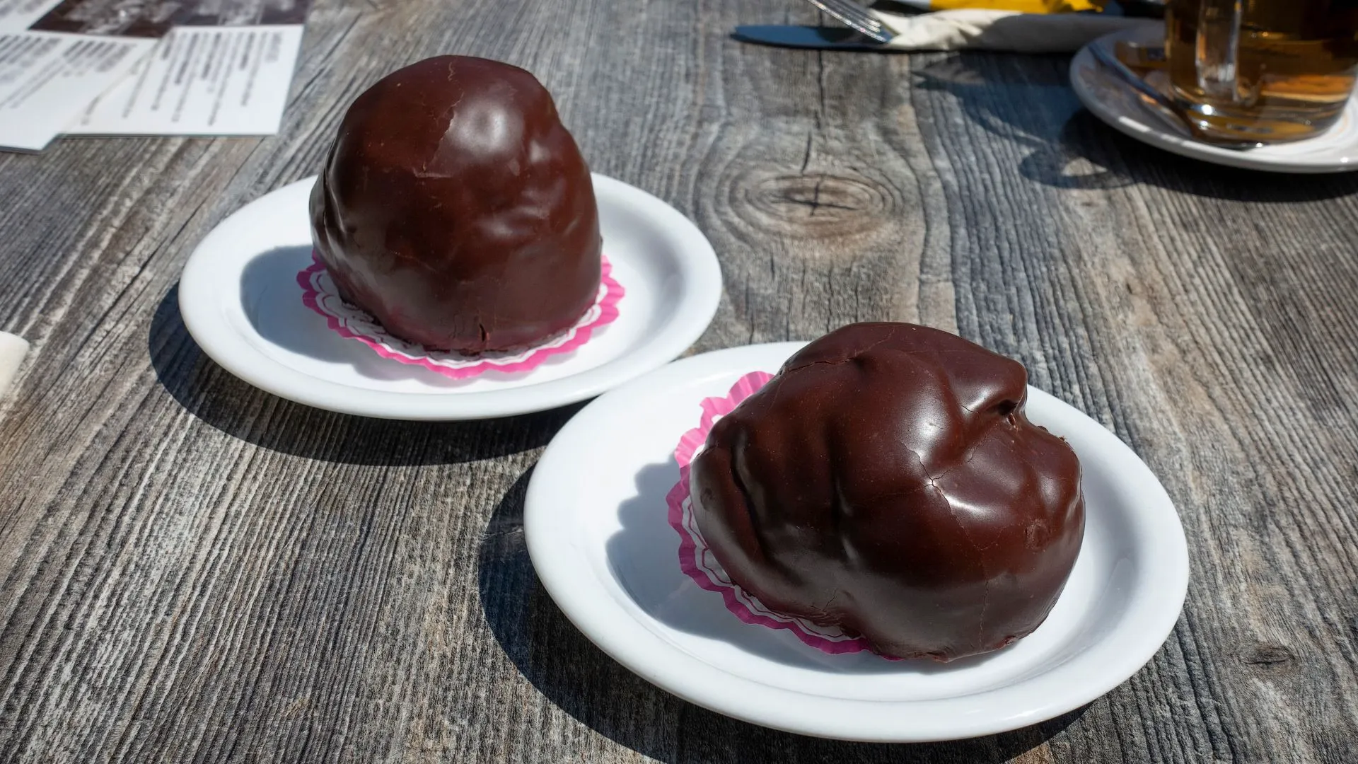 Two large chocolate-coated cream puffs on small white plates with pink doilies on a weathered wooden outdoor table
