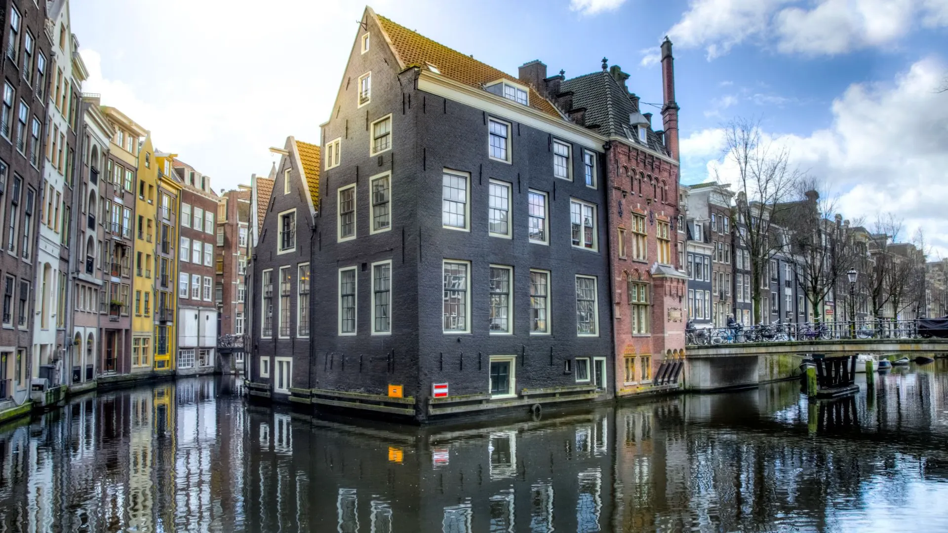 Historic Amsterdam canal houses on a corner where two canals meet, dark brick gabled buildings reflected in calm water under a blue sky