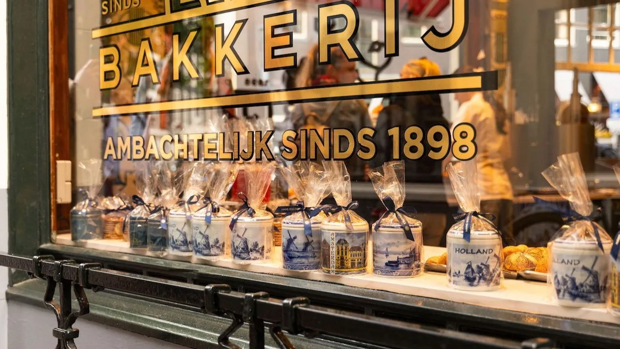 Amsterdam bakery window with gold lettering reading Ambachtelijk Sinds 1898 and a row of Delft blue jars wrapped in cellophane