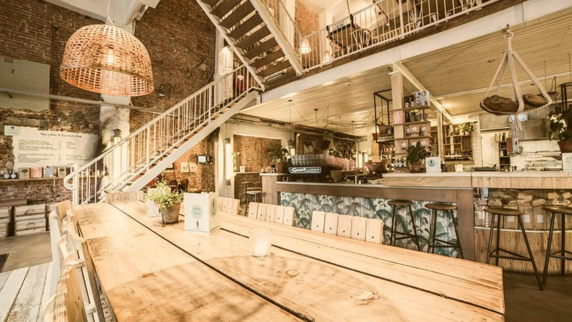 Spacious cafe interior with a long wooden communal table, white staircase, exposed brick walls and rattan lampshades