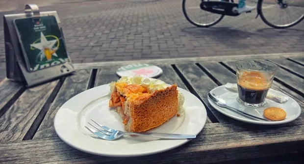 Slice of Dutch apple pie with whipped cream and an espresso on a wooden terrace table, a bicycle passing behind