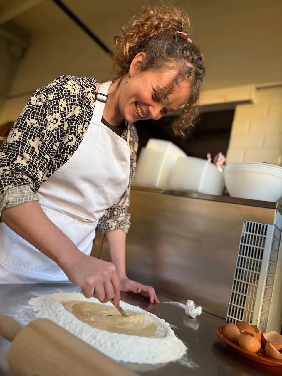 Smiling woman with curly hair in a white apron stirring eggs into a flour well on a steel counter, fresh eggs beside her