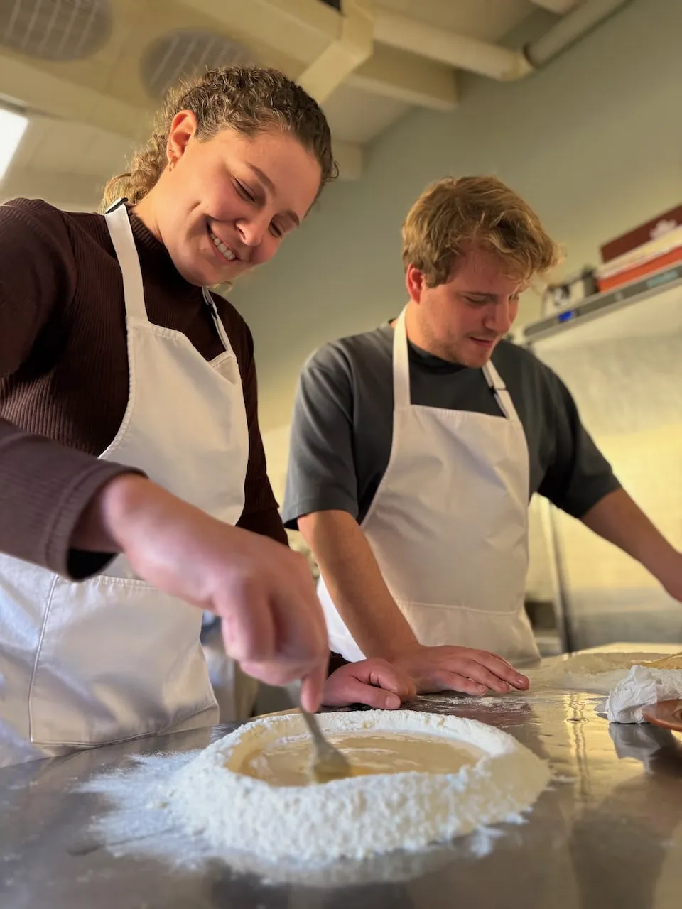 Young woman and man in white aprons mixing dough in a flour well on a stainless steel counter during a cooking class