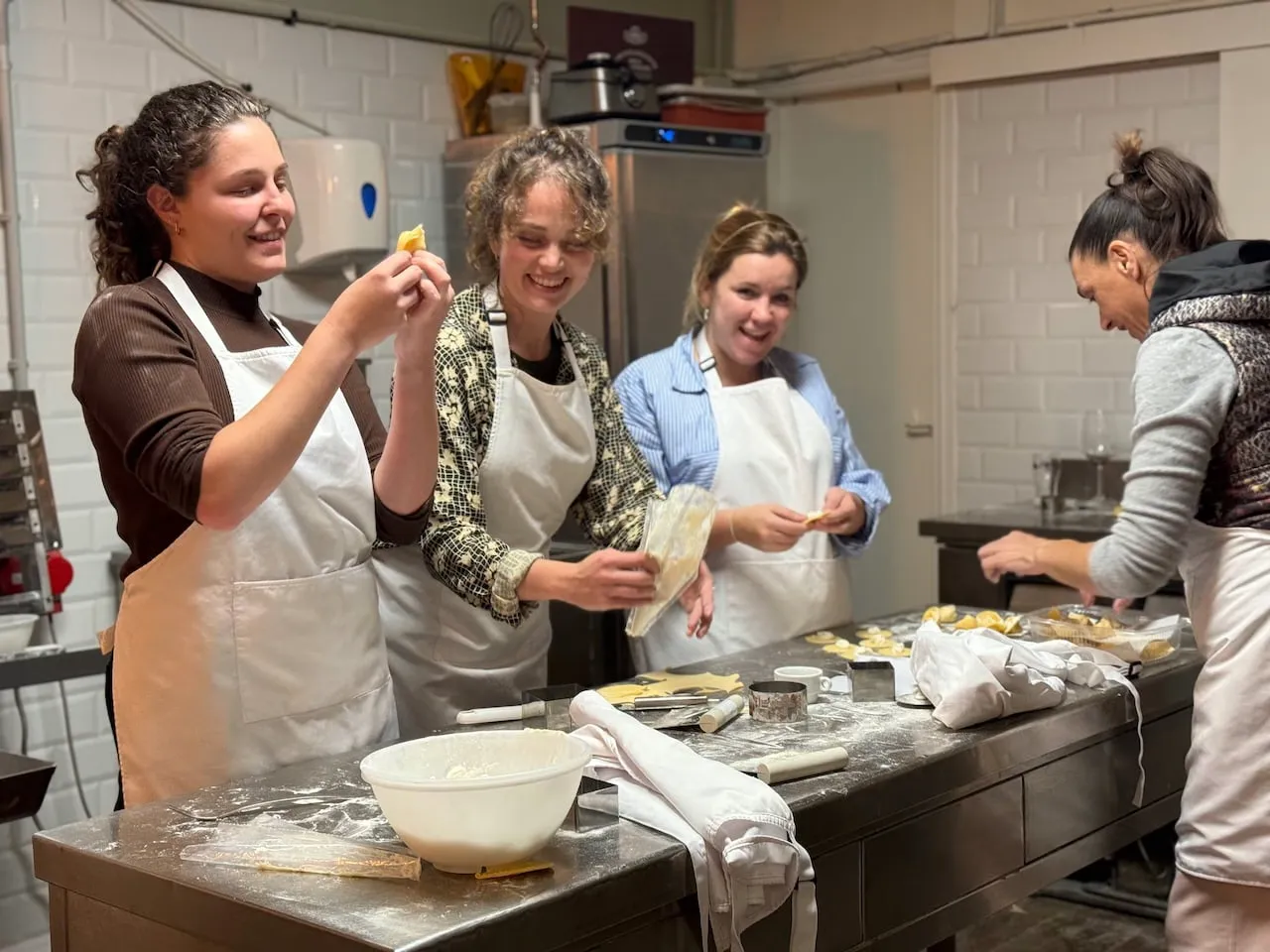 Four women in aprons laughing and tasting fresh dough creations around a steel worktable in a tiled kitchen