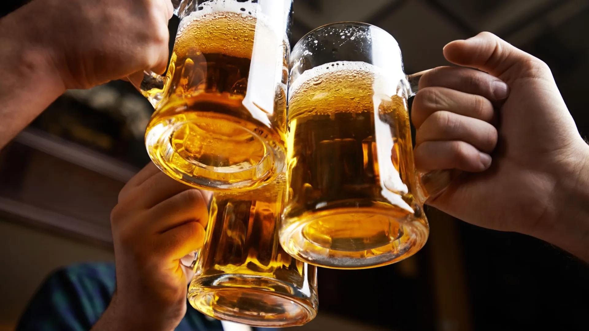 Three hands clinking full beer mugs together over a dark bar table