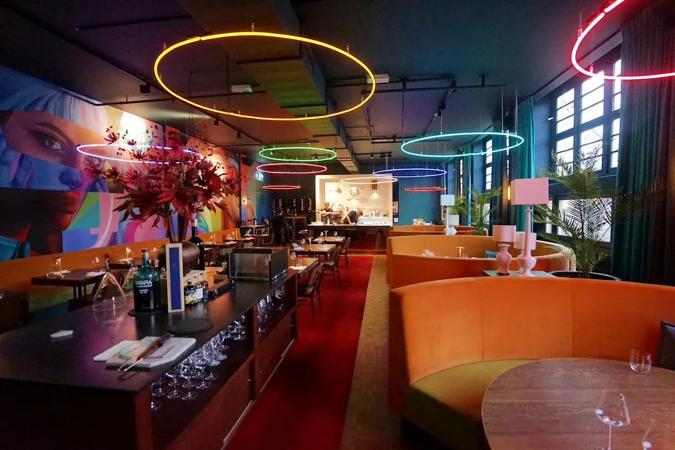 Modern restaurant interior with colorful neon light rings hanging from a dark ceiling, orange booths and a bold mural