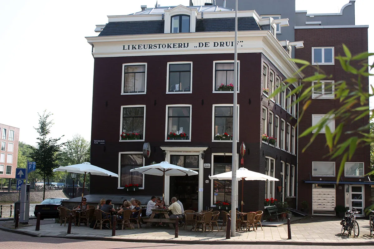 Historic dark brick corner building with Likeurstokerij De Druif lettering, white parasols and people on the terrace below