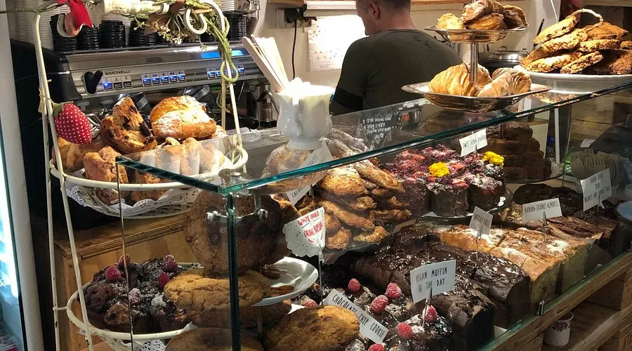 Glass bakery counter packed with cakes, scones, brownies and muffins, with a barista at the espresso machine behind