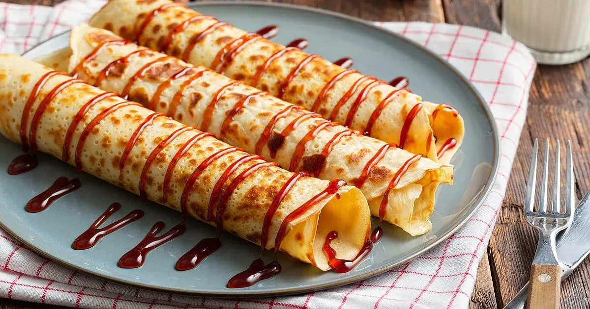 Three rolled thin pancakes drizzled with dark syrup on a grey plate over a red checked cloth