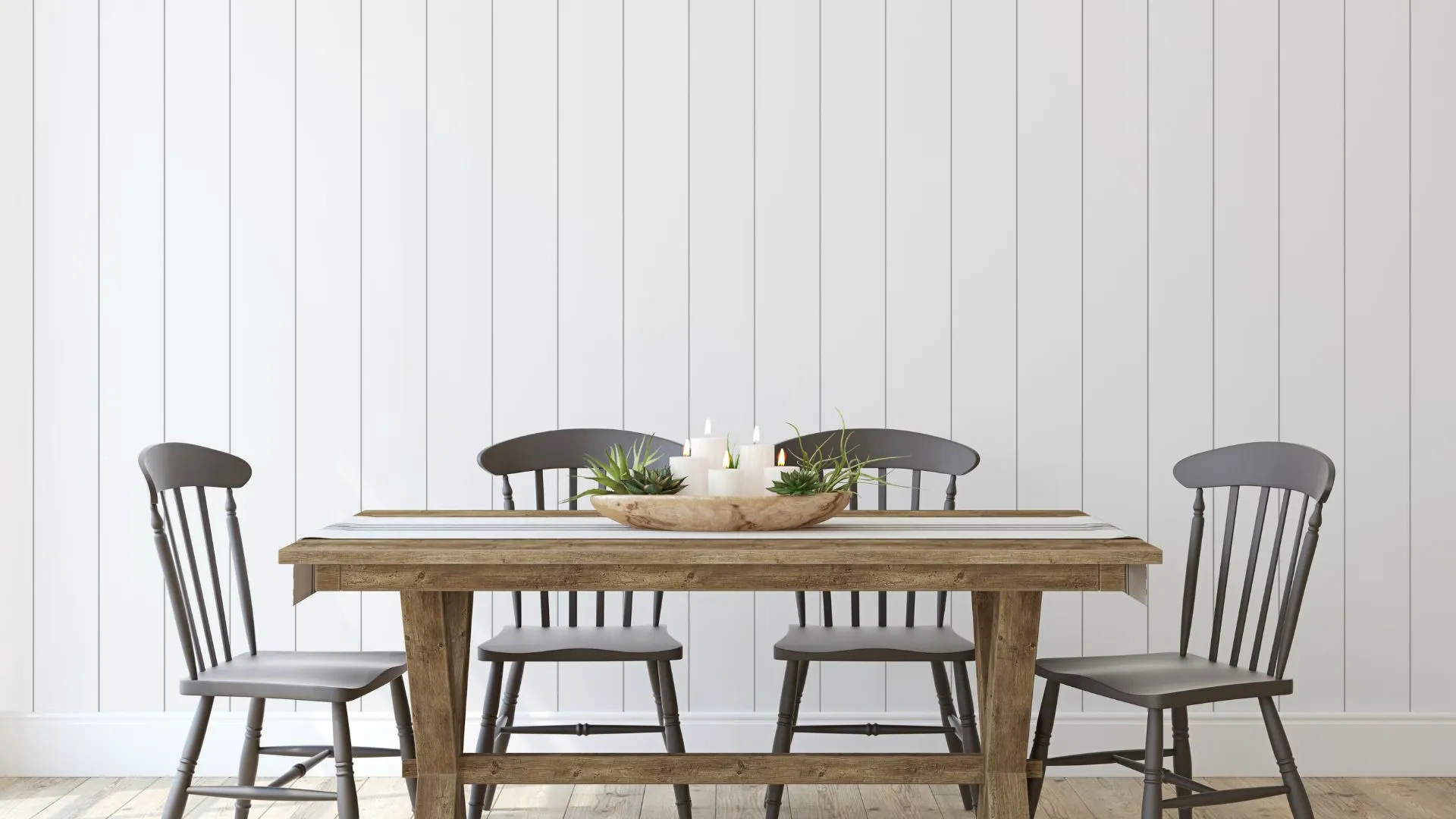Rustic wooden dining table with four grey chairs and a centerpiece of candles and plants against a white panel wall