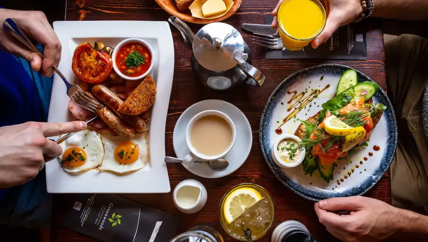 Overhead view of a cafe breakfast with fried eggs, sausages, grilled tomato, salmon toast, tea and orange juice on a dark wood table