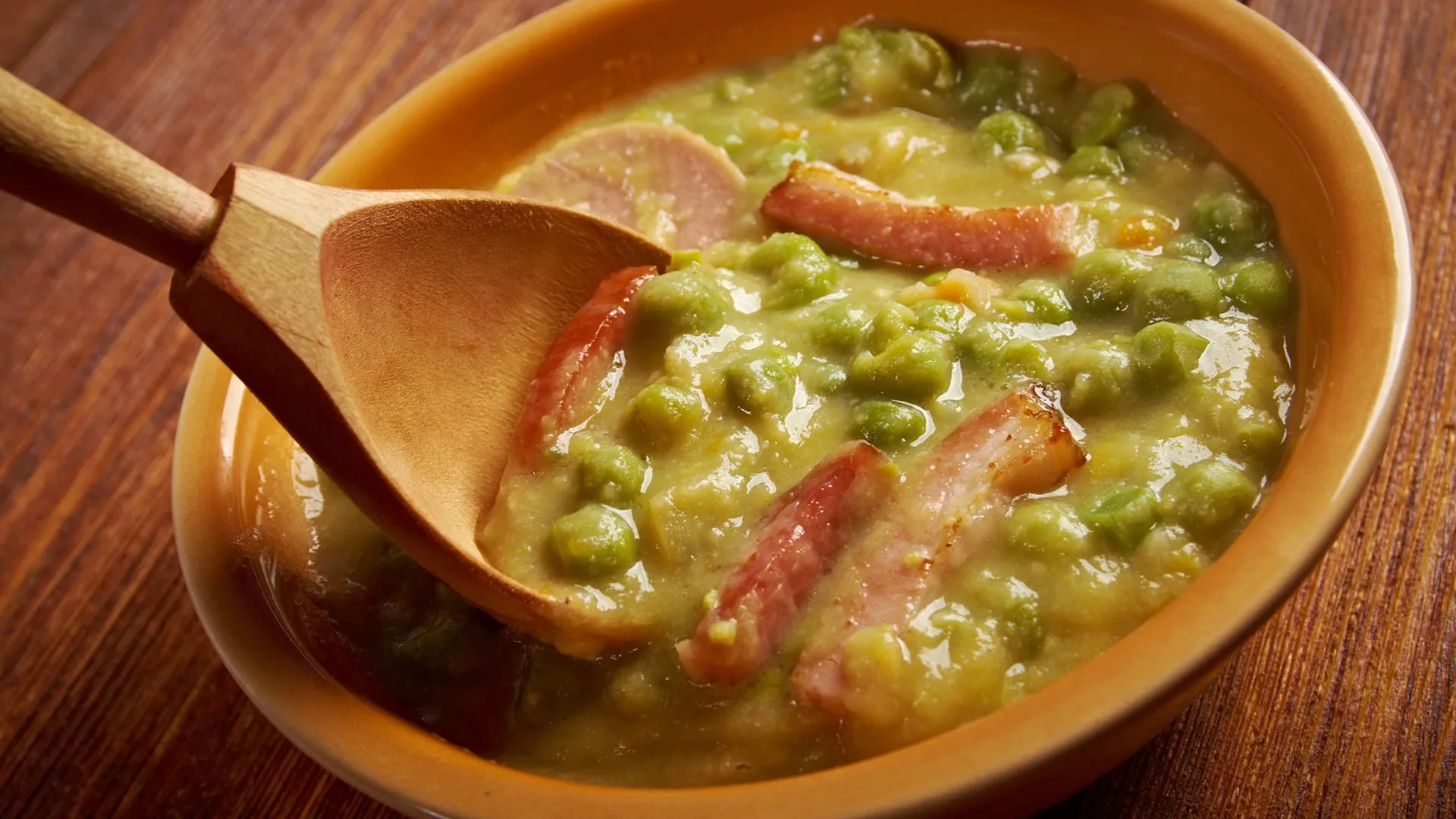 Thick green pea soup with slices of smoked sausage in an orange bowl, stirred with a wooden spoon