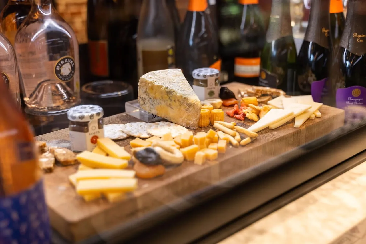Wooden board behind glass with wedges of blue and aged cheese, crackers and snacks, with spirit and wine bottles behind