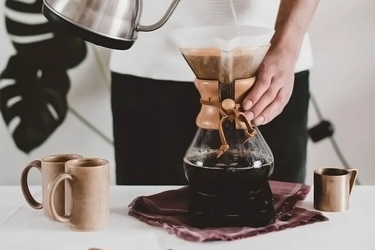 Hot water being poured from a gooseneck kettle into a glass pour-over coffee brewer, with ceramic mugs on the table