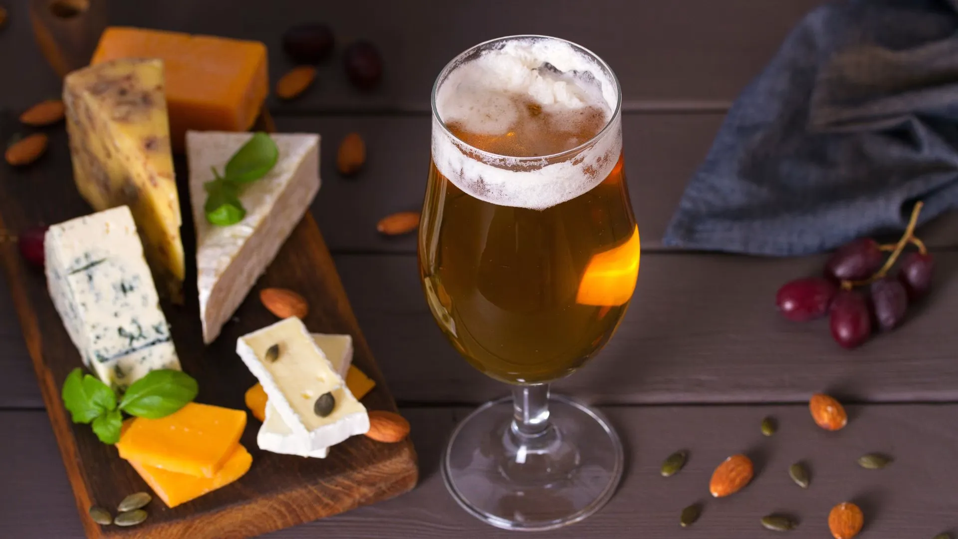 Glass of amber craft beer with thick foam next to a wooden board of assorted cheeses, almonds and grapes