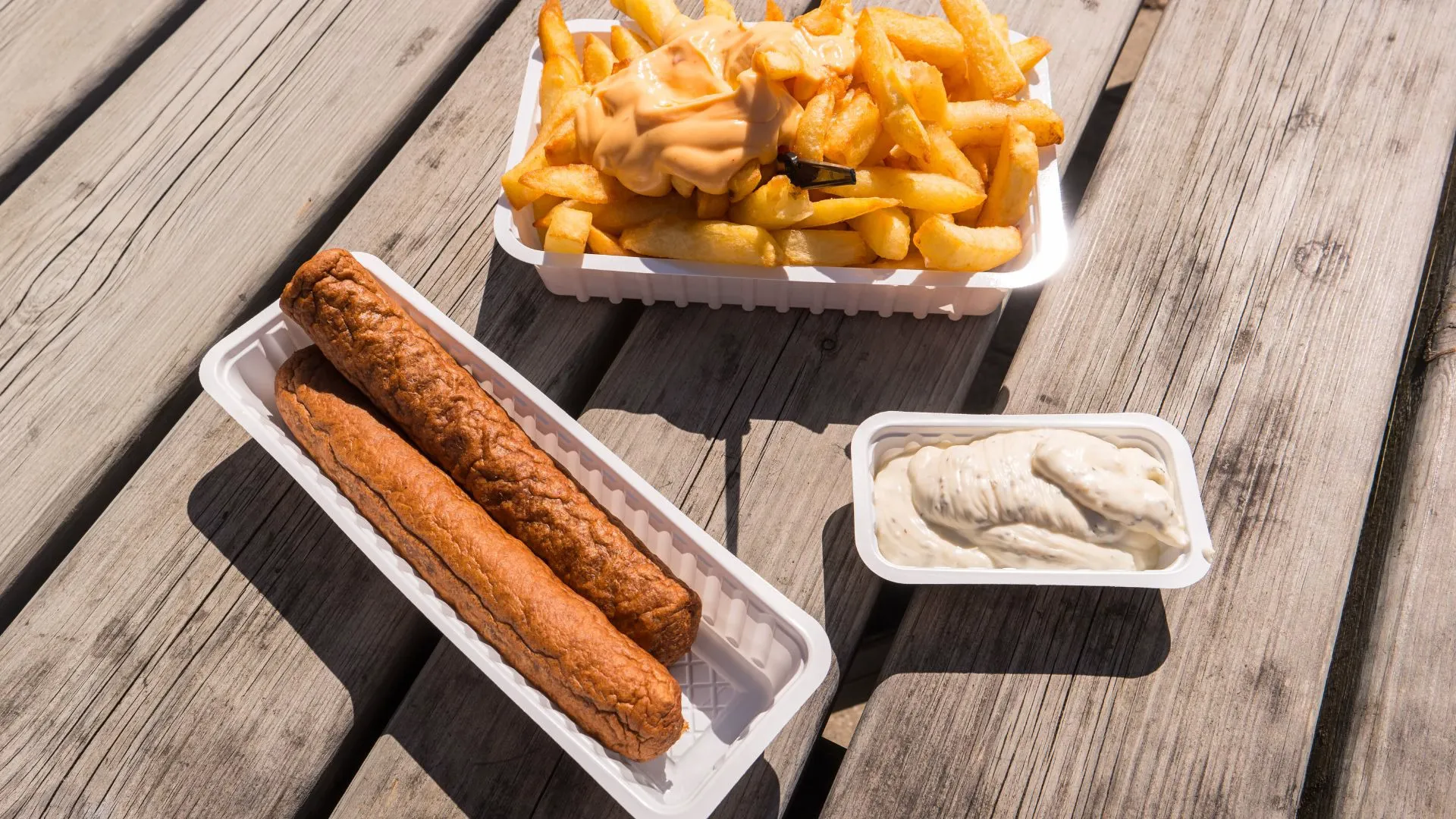 Two frikandellen in a paper tray next to fries with sauce and a tub of mayonnaise on a wooden picnic table