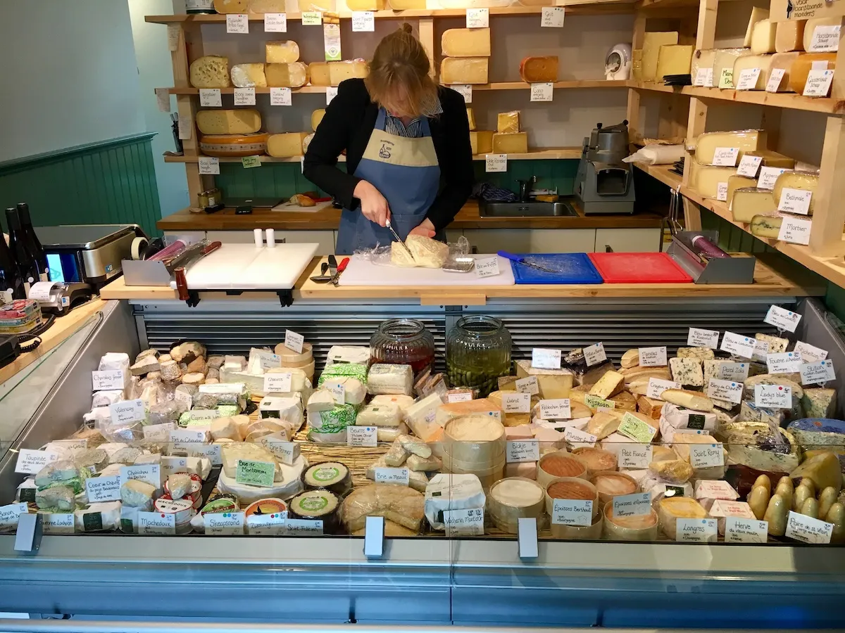 Cheesemonger cutting a wheel of cheese behind a counter packed with labeled cheeses at a fromagerie