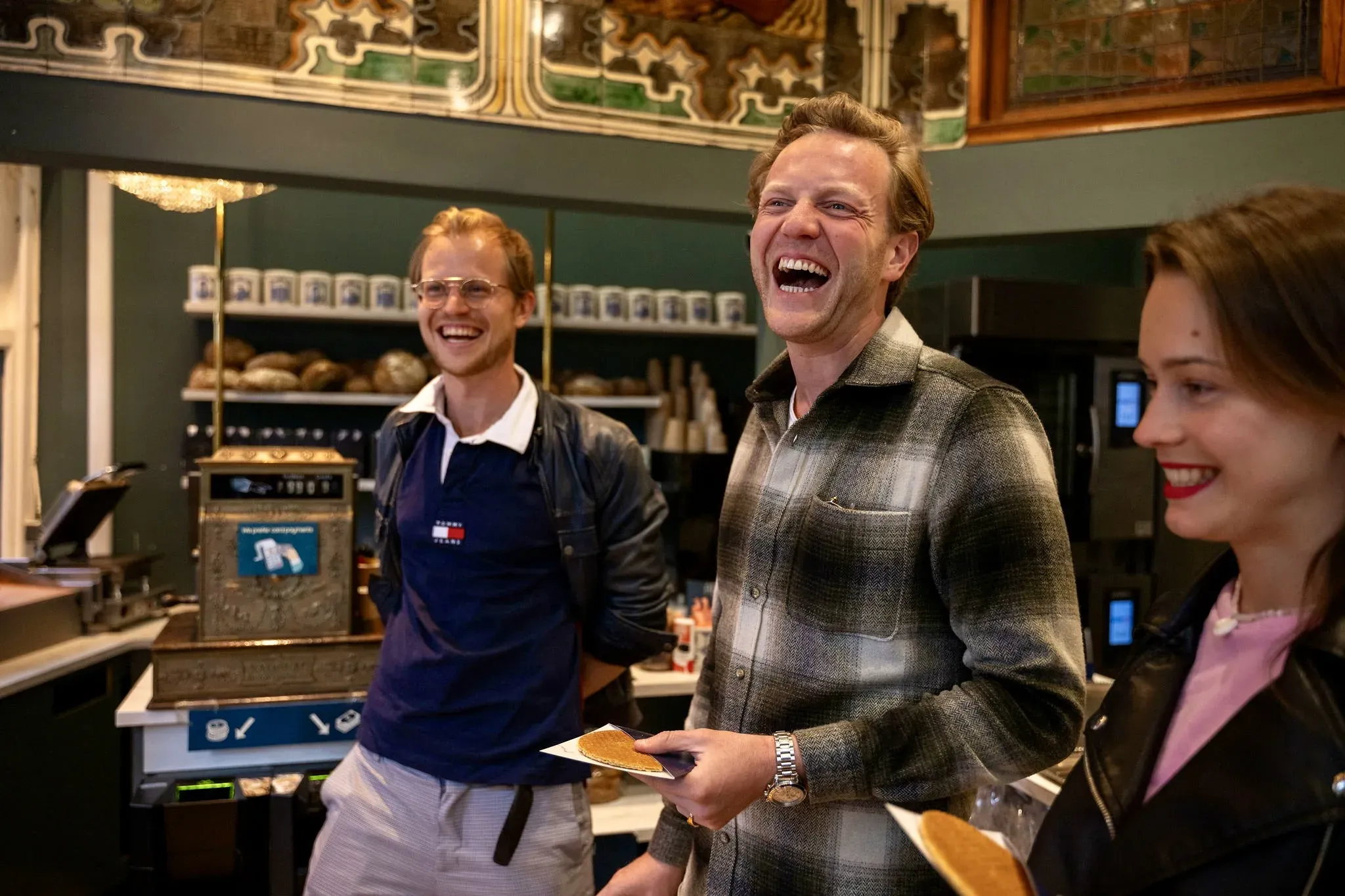Three guests laughing while holding fresh stroopwafels inside a historic Amsterdam bakery with tiled walls