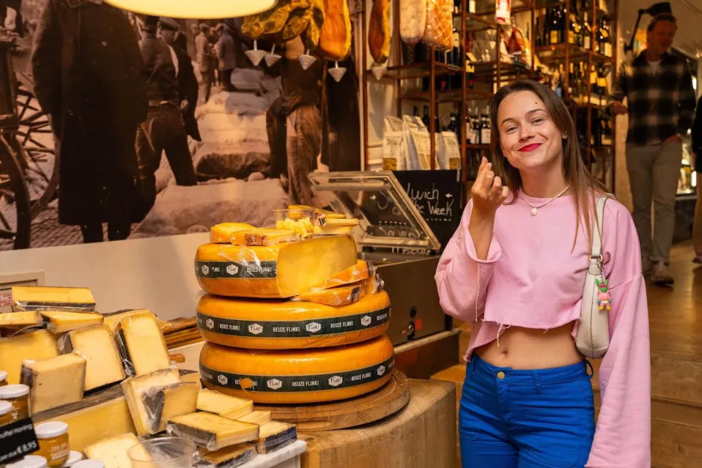 Woman in a pink sweater makes a chef's kiss gesture next to a stack of large Gouda cheese wheels in an Amsterdam cheese shop