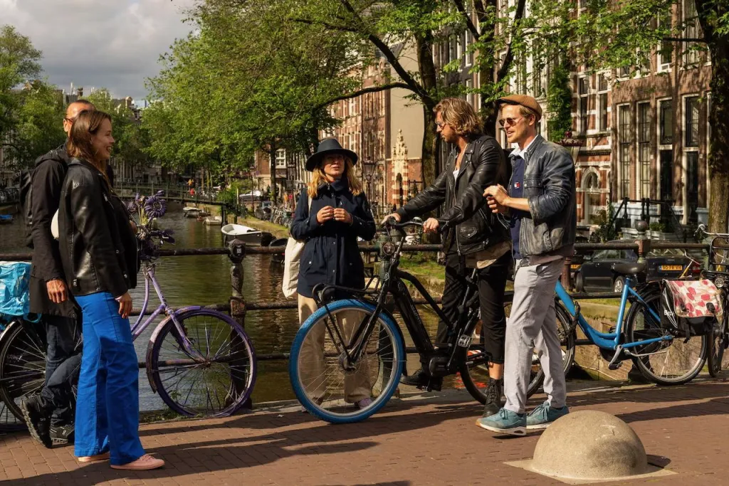 Group of five with bicycles talks together on a sunlit canal bridge in Amsterdam, old brick houses and trees behind them