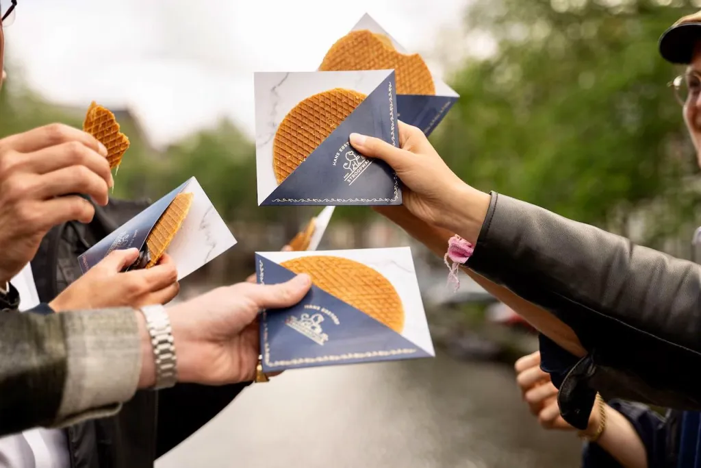 Four hands hold fresh stroopwafels in blue paper sleeves together in a toast above an Amsterdam canal