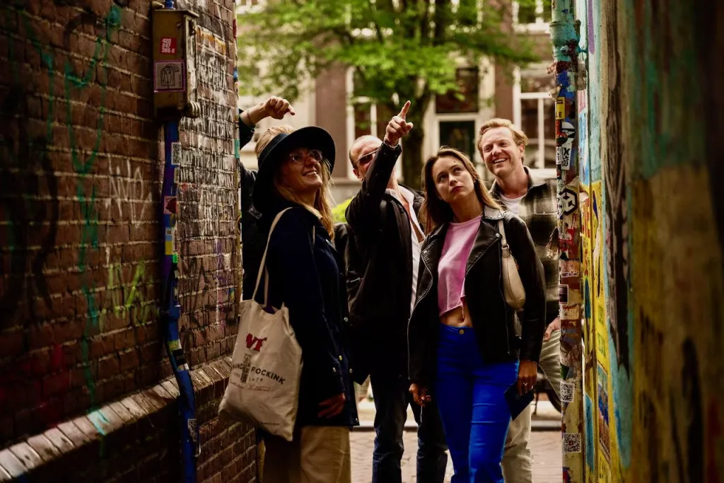 Tour guide points upward as a small group looks up in a narrow Amsterdam alley covered in graffiti and stickers