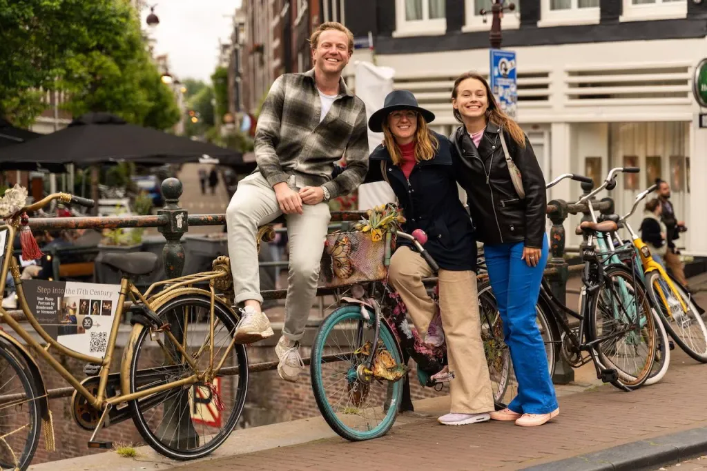 Three smiling friends pose with vintage bicycles on a canal bridge railing in Amsterdam