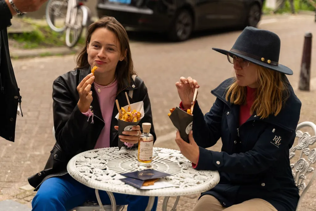 Two women eat loaded fries from paper cones at a white cast-iron terrace table on an Amsterdam street
