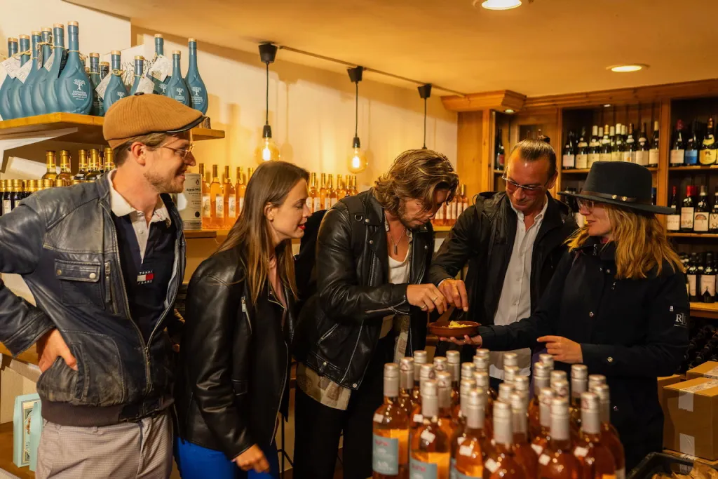 Five people gather around a small dish to taste something in a wine shop lined with bottles on wooden shelves