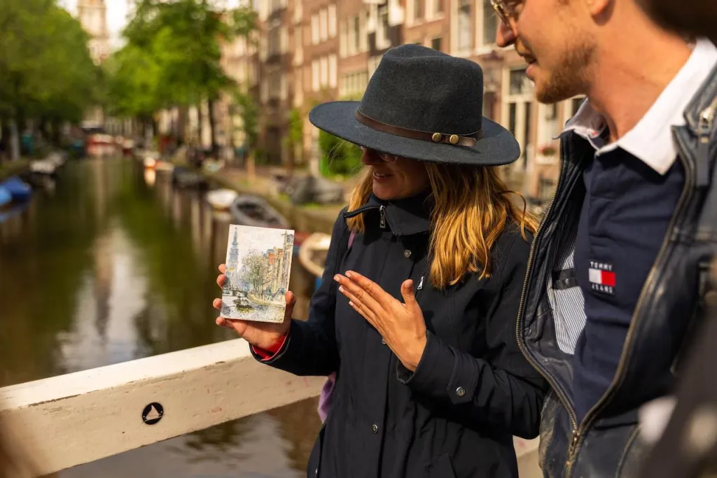 Woman in a wide-brimmed hat holds up a small painting of an Amsterdam canal scene while standing on a bridge over that canal